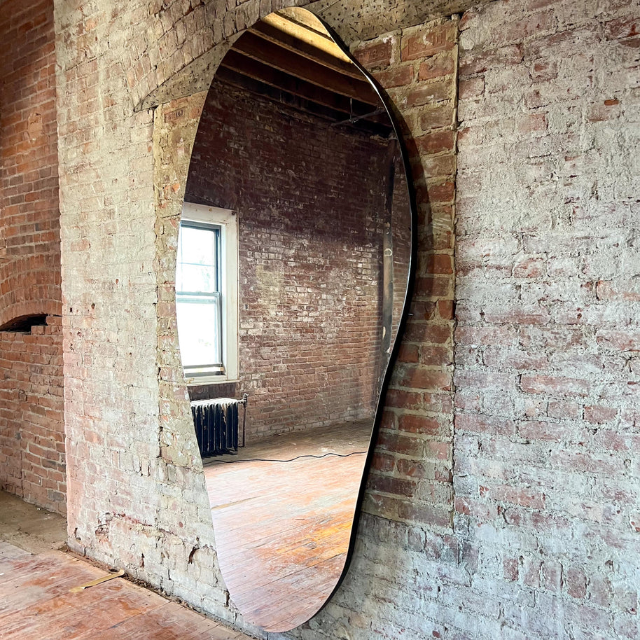 Large asymmetrical oval mirror mounted against a brick wall.