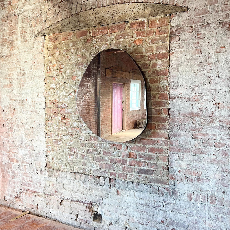 Small asymmetrical mirror on a brick wall with a view of a pink door.
