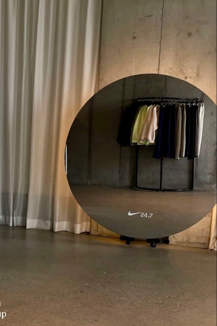 Room with a round shatterproof mirror with vinyl lettering of nike logo reflecting a closet and clothes, with a curtain in the background.